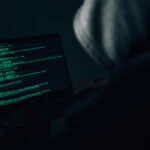 Behind the scenes of an anonymous hacker wearing a hoodie over his head in the dark hacking into a victim's financial system. Data thief, internet fraud, darknet and cyber security concept.