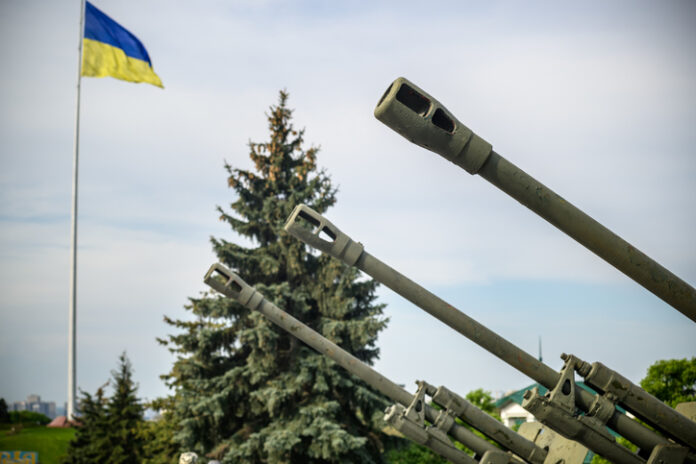 Groups of ancient military guns against the background of the State Flag of Ukraine. Muzzle brake of an artillery gun. Sunny morning sky. Call to stop violence concept. Ukrainian flag.